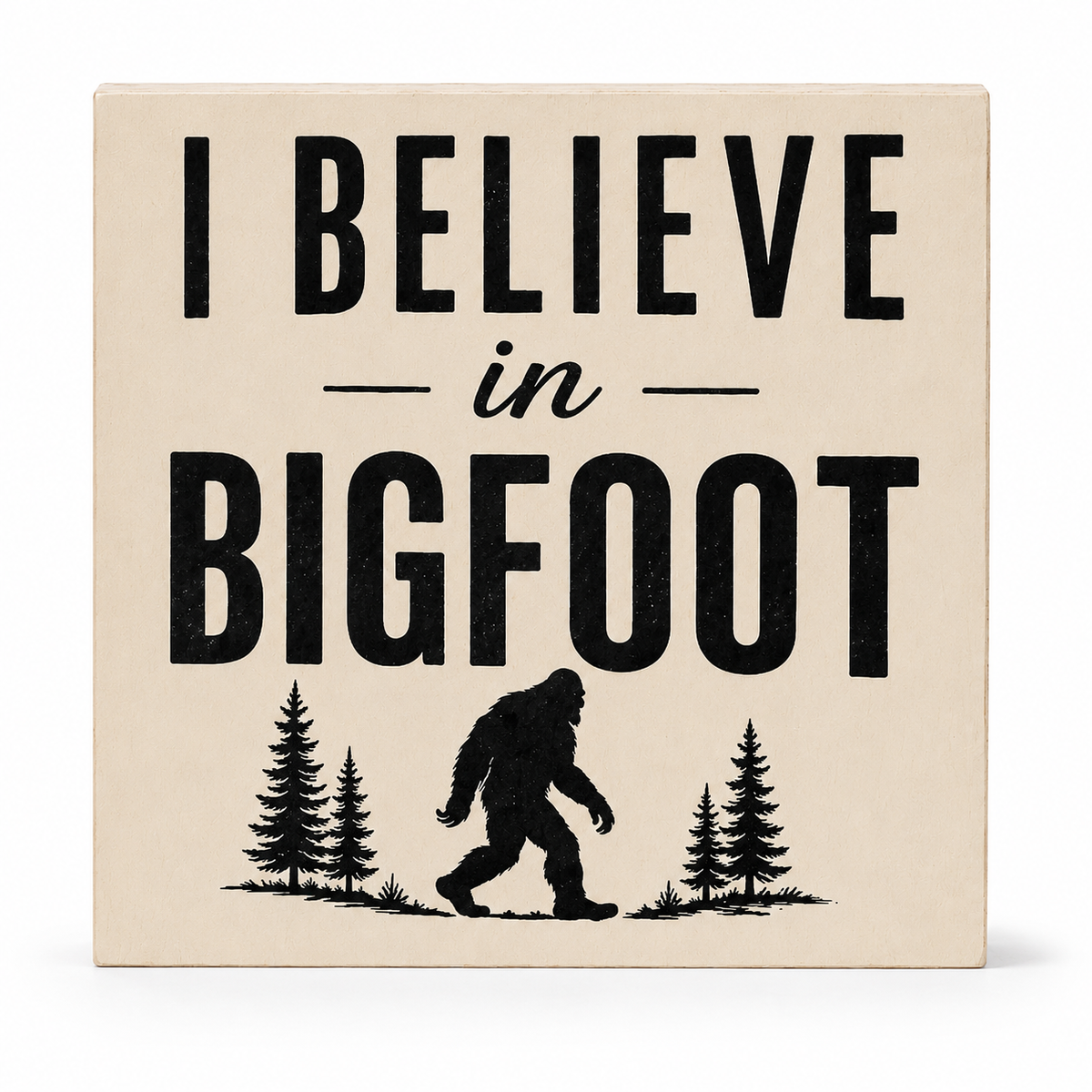 A square beige decorative sign with black text reading 'I BELIEVE in BIGFOOT' and a black silhouette of a Bigfoot figure walking between pine trees at the bottom, shown on a clean white background.