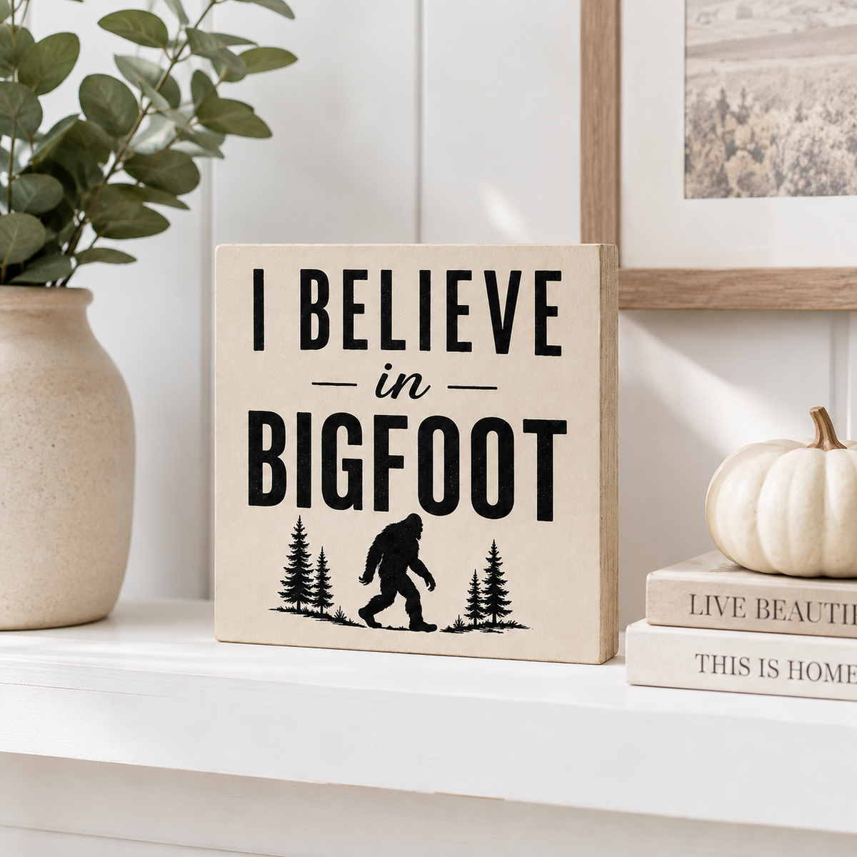 A square beige wooden sign with black text reading 'I BELIEVE in BIGFOOT' and a black silhouette of Bigfoot among pine trees, styled on a white shelf with a vase, framed art, books, and a small pumpkin.