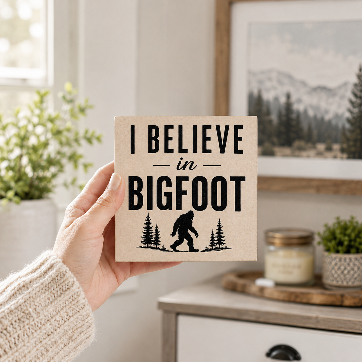 A hand wearing a cream knit sweater holds a square wooden sign that reads 'I BELIEVE in BIGFOOT' with a black silhouette of a Bigfoot figure and pine trees. The background shows a cozy, softly blurred indoor setting with a window, green plants, and a framed mountain landscape print.
