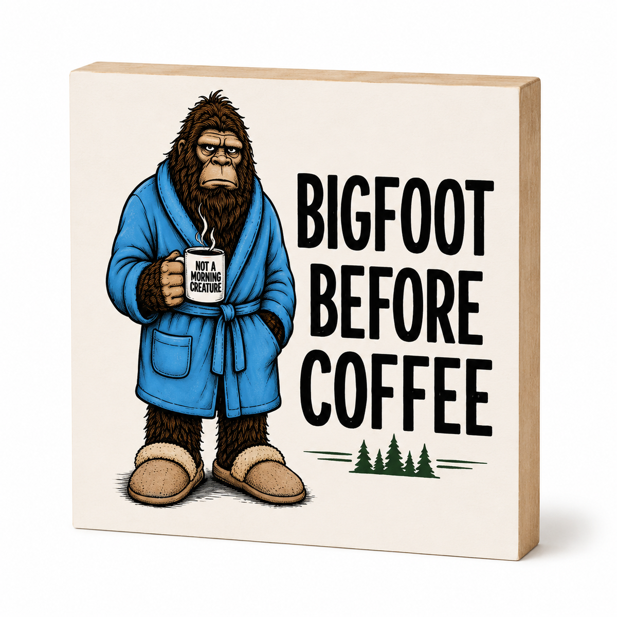 A square decorative sign or block with a printed illustration of Bigfoot in a blue robe holding a mug, next to the text 'BIGFOOT BEFORE COFFEE' and small pine trees, shown on a clean white background.