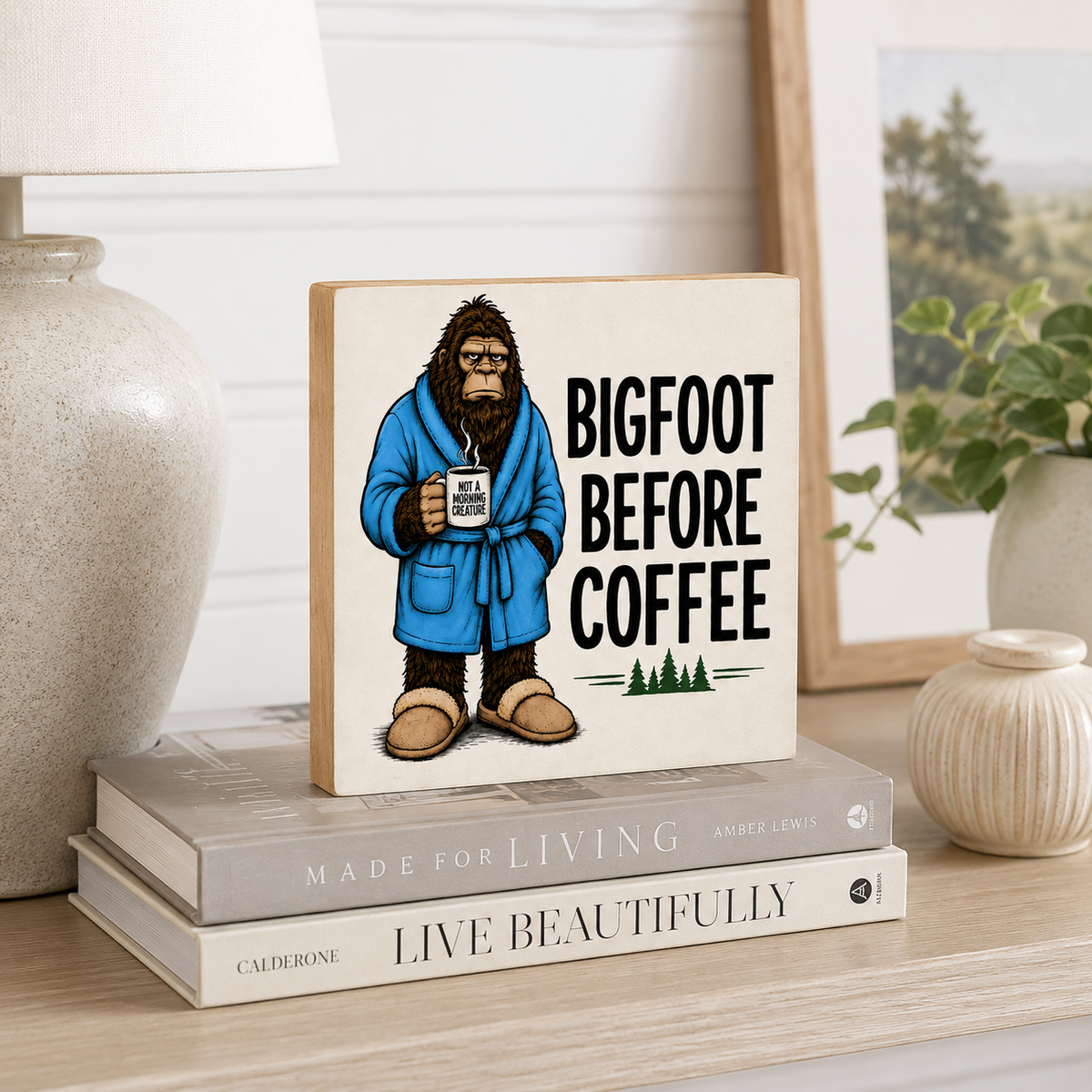 A square decorative sign with a wood-grain border stands on stacked books on a shelf. The sign shows an illustrated Bigfoot wearing a blue robe and holding a mug, with the text 'BIGFOOT BEFORE COFFEE.