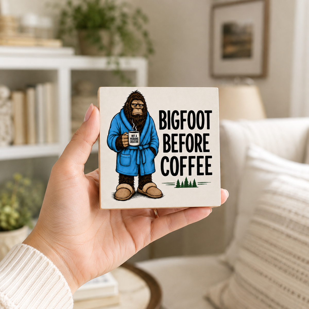 A hand holds a square ceramic coaster featuring a cartoon Bigfoot in a blue robe holding a mug with the text 'BIGFOOT BEFORE COFFEE'. The background shows a cozy, softly blurred living room interior with a sofa, lamp, and bookshelf.