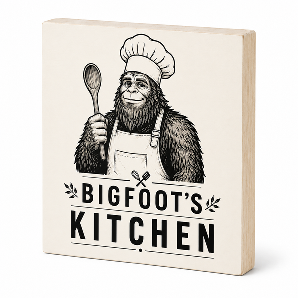 A square wooden decorative sign with a light natural wood edge and a cream face, printed with a black illustration of Bigfoot wearing a chef hat and apron while holding a spoon, above the text 'BIGFOOT'S KITCHEN'.