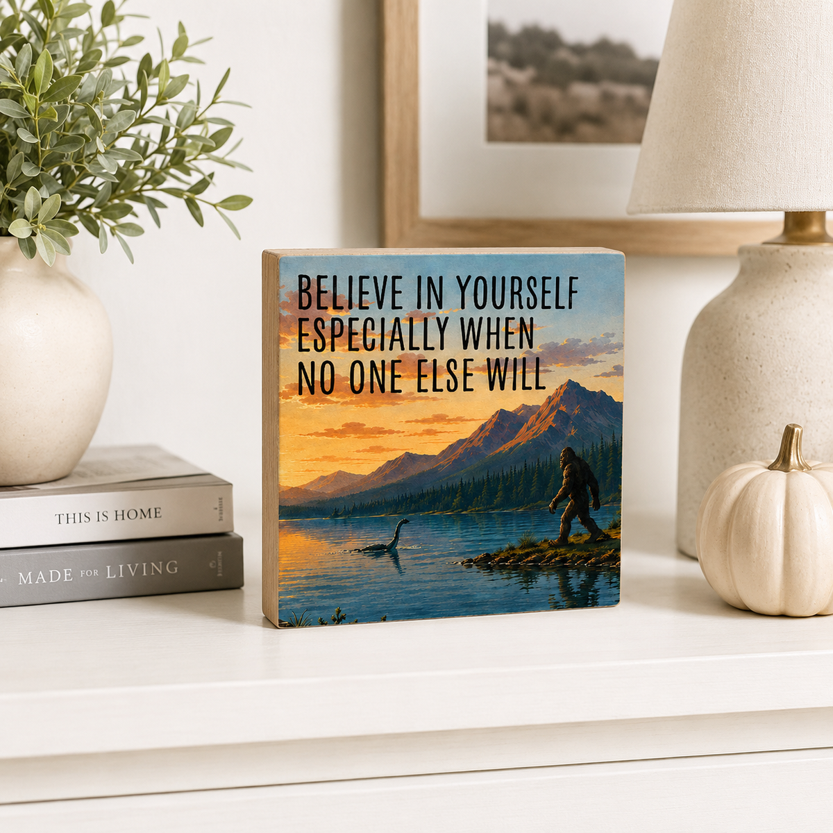 A square wooden sign with a scenic mountain lake sunset print and the text 'BELIEVE IN YOURSELF ESPECIALLY WHEN NO ONE ELSE WILL' stands on a white shelf beside a vase, books, a lamp, and a small pumpkin.