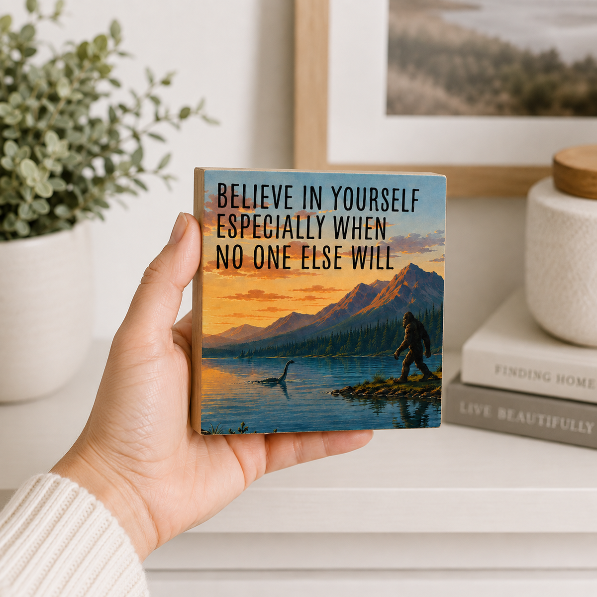 A hand holds a square wooden block or coaster featuring a scenic mountain lake sunset illustration and the text 'BELIEVE IN YOURSELF ESPECIALLY WHEN NO ONE ELSE WILL'. The background shows a softly blurred home interior with a plant, framed art, books, and a ceramic jar.