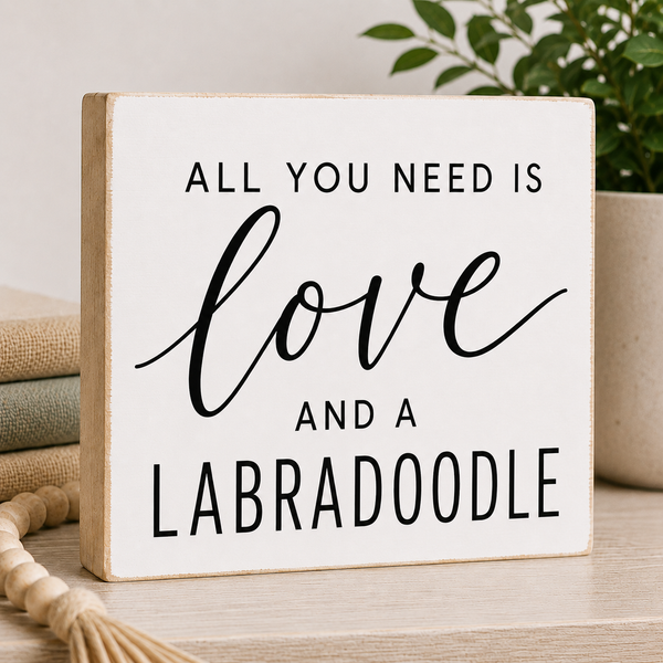 A square white decorative sign with a light natural wood border, featuring the text 'ALL YOU NEED IS love AND A LABRADOODLE' in black lettering. The sign is styled on a light wood tabletop with a tassel rope, stacked books, and a potted plant in the background.