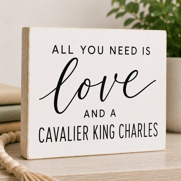 A rectangular white wooden sign with black text reading 'ALL YOU NEED IS love AND A CAVALIER KING CHARLES' displayed on a light wood surface with stacked books, a rope, and a potted plant in the background.