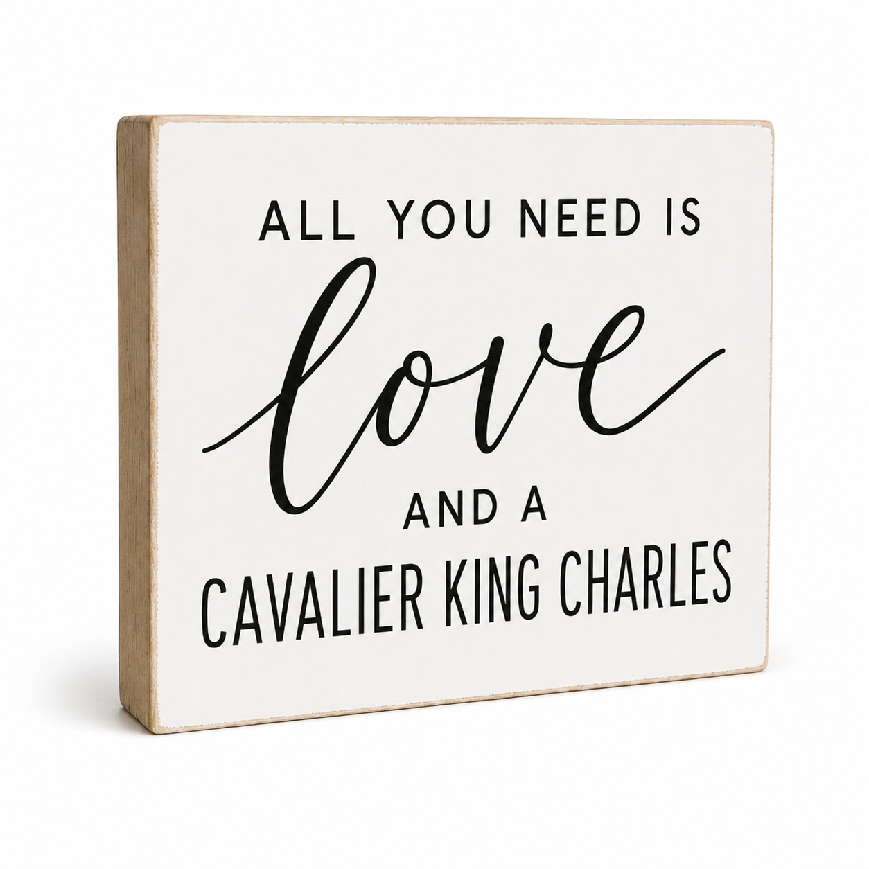A square decorative wooden sign with a white face and natural wood edges, printed with the phrase 'ALL YOU NEED IS love AND A CAVALIER KING CHARLES' in black lettering, shown on a light gray background.