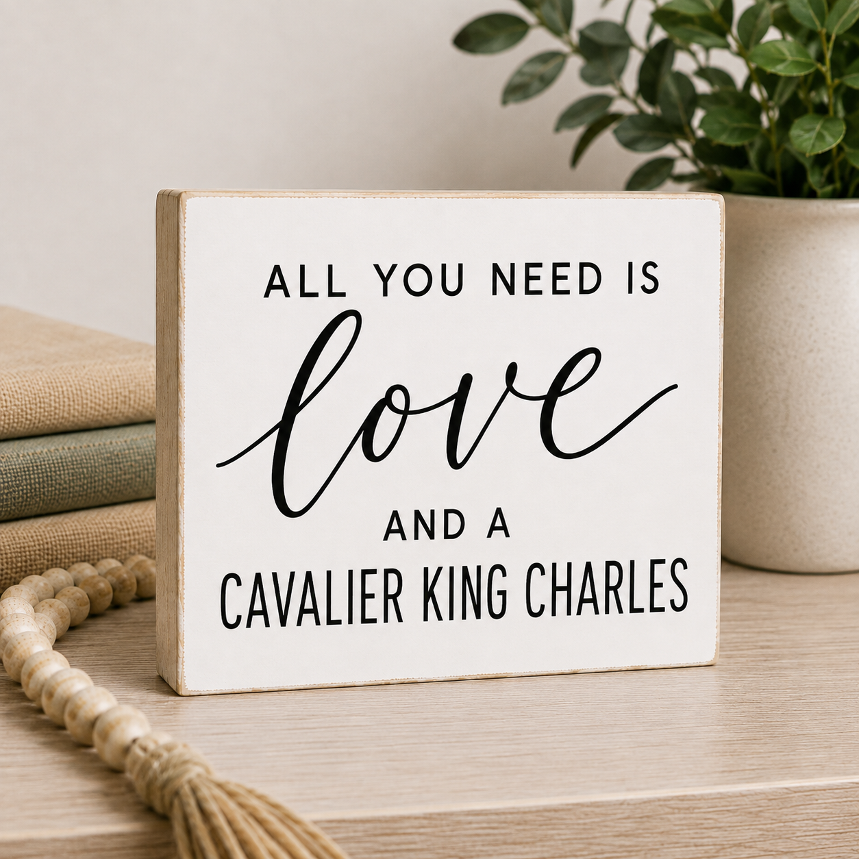 A square white decorative sign with a light wood border and black text reading 'ALL YOU NEED IS love AND A CAVALIER KING CHARLES,' styled on a tabletop with a plant, books, and a tassel in the background.