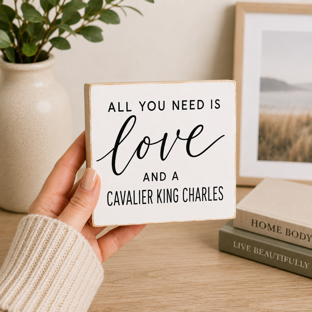 A hand wearing a cream knit sweater holds a square white wooden sign with black text that reads, 'ALL YOU NEED IS love AND A CAVALIER KING CHARLES.' The background shows a softly styled interior with a vase, framed picture, and stacked books on a wooden surface.