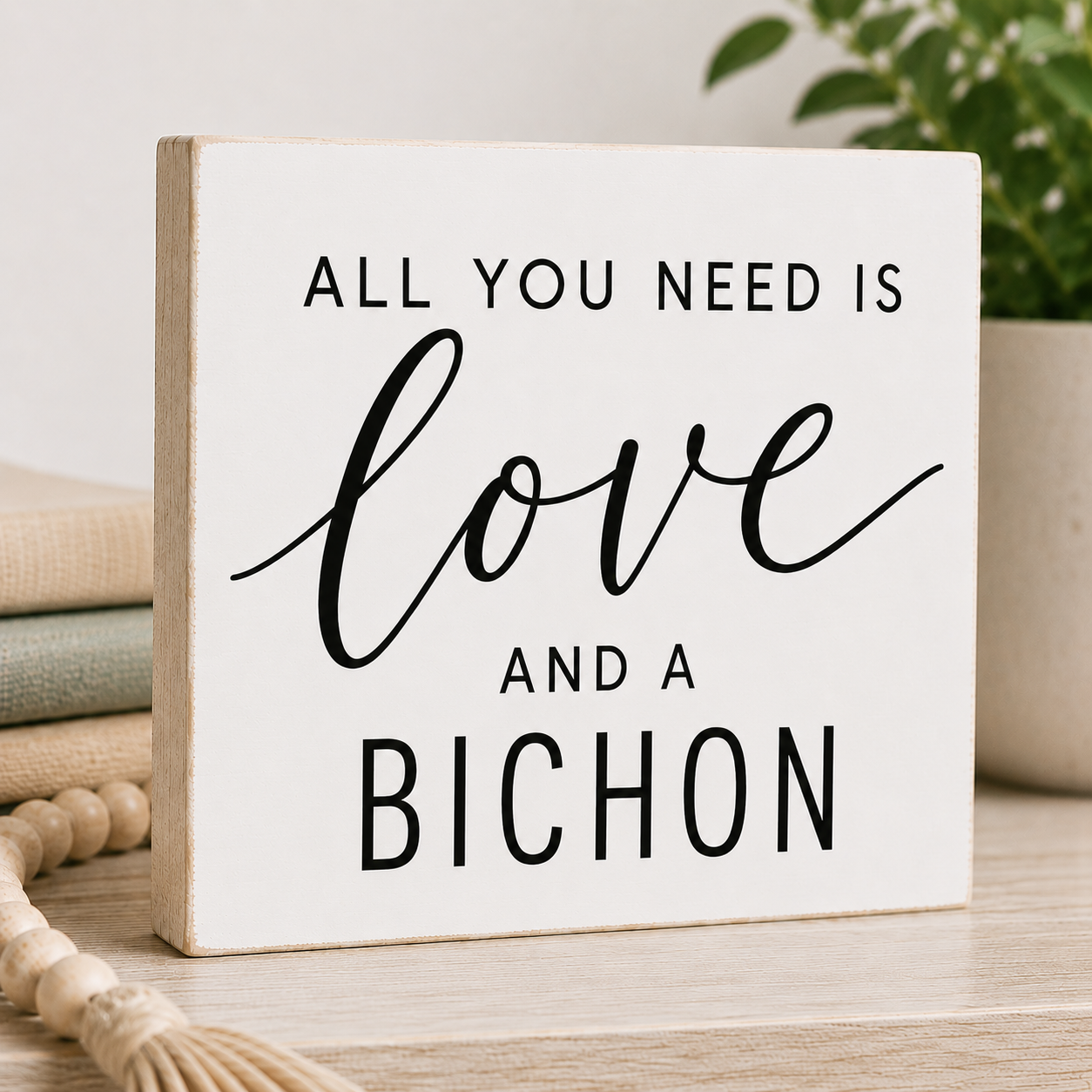 A square white decorative sign with a light wood edge, printed with the phrase 'ALL YOU NEED IS love AND A BICHON' in black lettering, displayed on a tabletop with stacked books, a plant, and a woven tassel in the background.