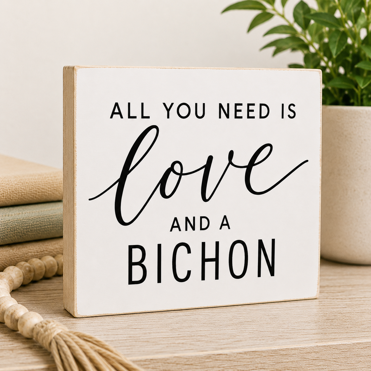 A square white decorative wooden sign with black text reading 'ALL YOU NEED IS love AND A BICHON' displayed on a light wood surface with stacked books, a plant, and a woven tassel nearby.