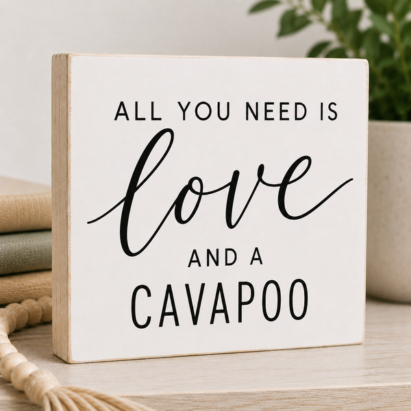 A square white decorative sign with black text reading 'ALL YOU NEED IS love AND A CAVAPOO' styled on a tabletop with books, a plant, and a rope in the background.