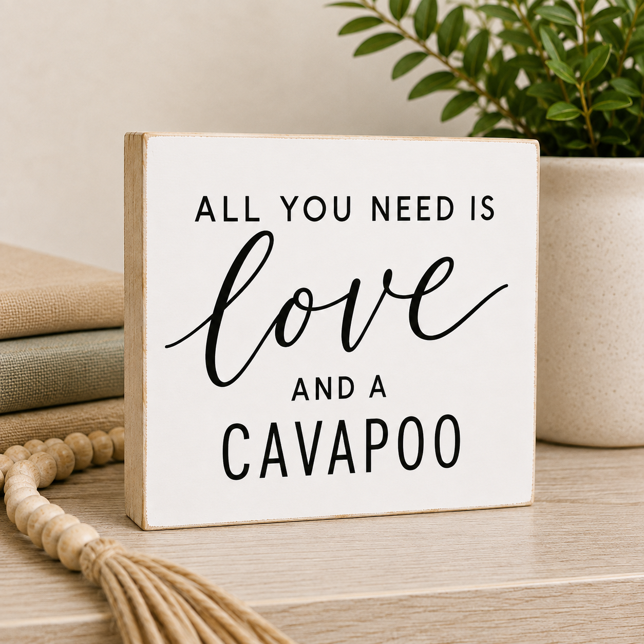 A square white decorative wooden sign with a natural wood edge and black text reading 'ALL YOU NEED IS love AND A CAVAPOO', styled on a tabletop with stacked books, a tassel, and a potted plant in the background.