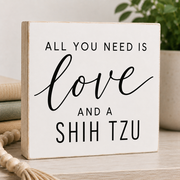 A square white wooden sign with a light natural wood edge, featuring the text 'ALL YOU NEED IS love AND A SHIH TZU' in black lettering. The sign is styled on a light wood surface with a neutral background, including stacked books, a decorative rope, and a potted plant.