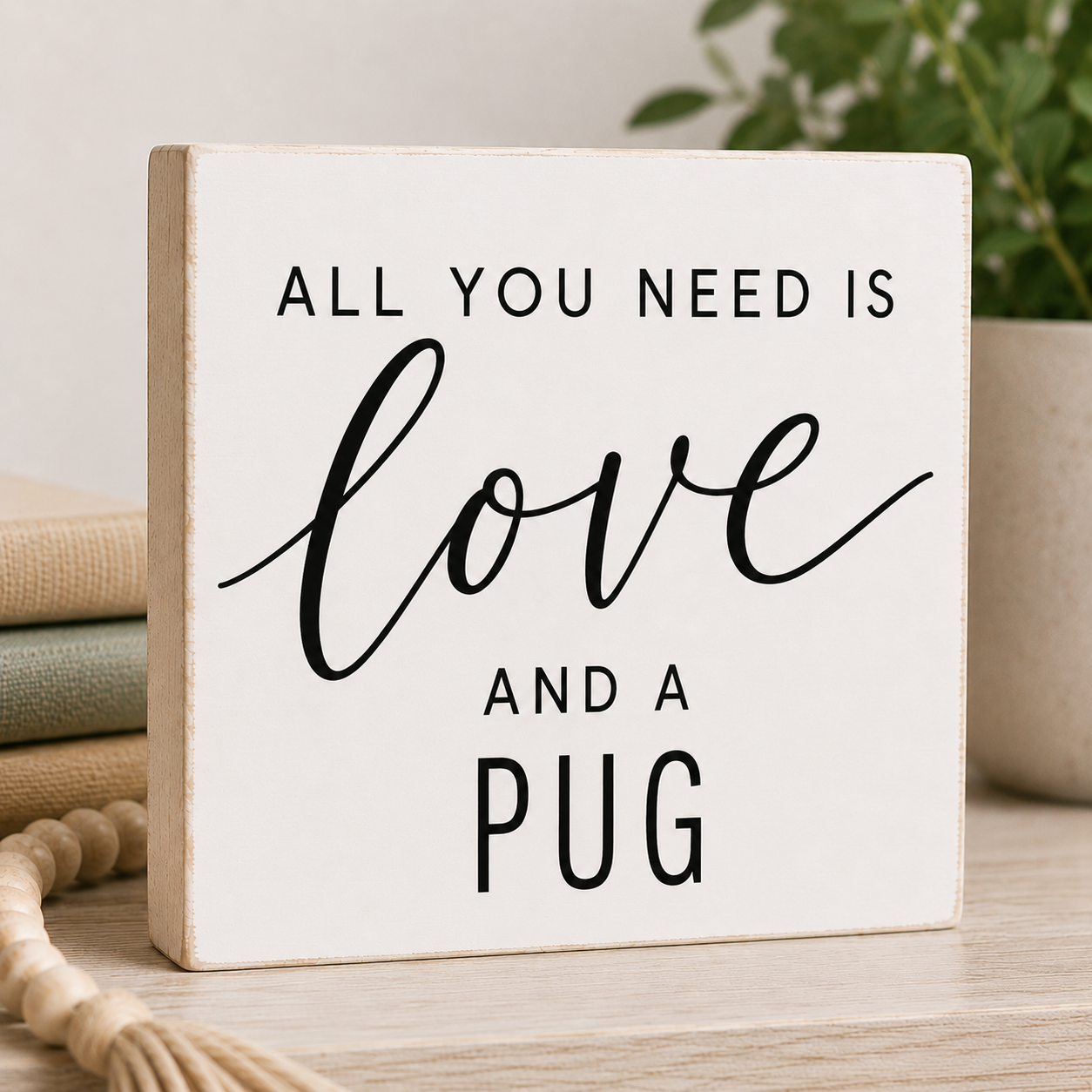 A square white wooden sign with black text reading 'ALL YOU NEED IS love AND A PUG' styled on a light wood surface with neutral decor in the background.
