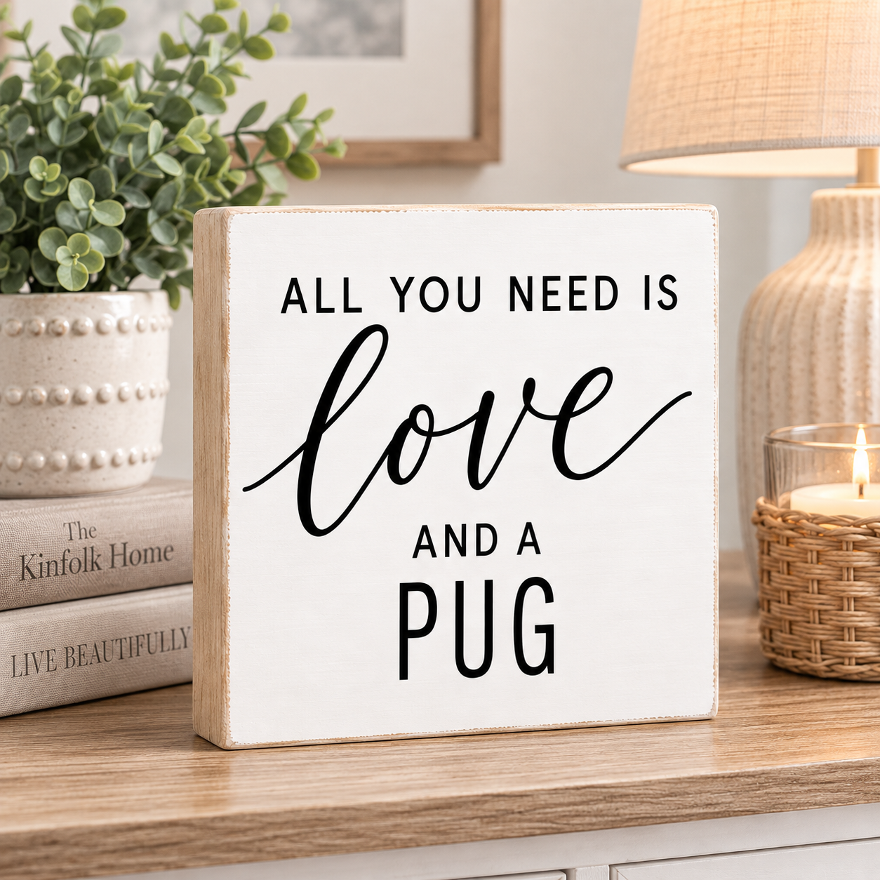 A square white decorative wooden sign with a light natural wood edge, featuring the text 'ALL YOU NEED IS love AND A PUG' in black lettering. The sign is styled on a wooden table in a cozy room setting with a potted plant, books, a lamp, and a lit candle in the background.