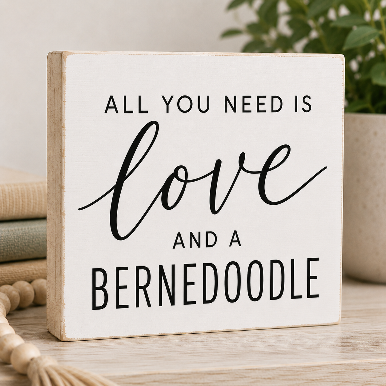 A square white decorative wooden sign with black text reading 'ALL YOU NEED IS love AND A BERNEDOODLE' displayed on a light wood surface with stacked books, a tassel, and a potted plant in the background.