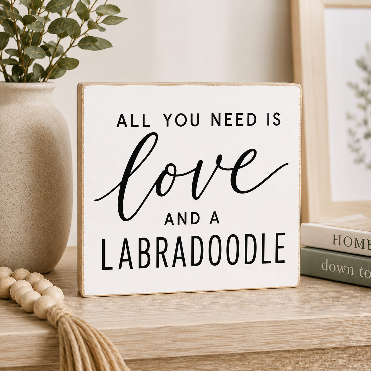 A square white decorative sign with a light wood border and black text reading 'ALL YOU NEED IS love AND A LABRADOODLE,' displayed on a wooden shelf beside a beige vase with greenery and stacked books.