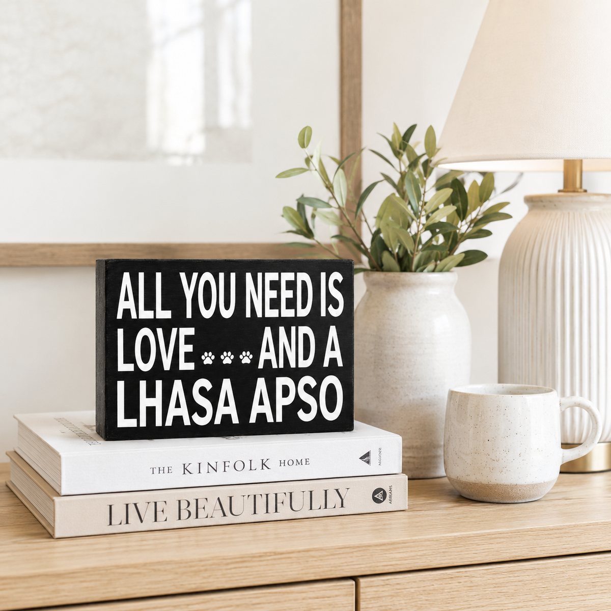 A black rectangular decorative sign with bold white text reading 'ALL YOU NEED IS LOVE *** AND A LHASA APSO' sits on top of two white books on a light wood console. A ceramic vase with greenery, a ribbed cream lamp, and a matching ceramic mug are arranged around it in a styled interior setting.