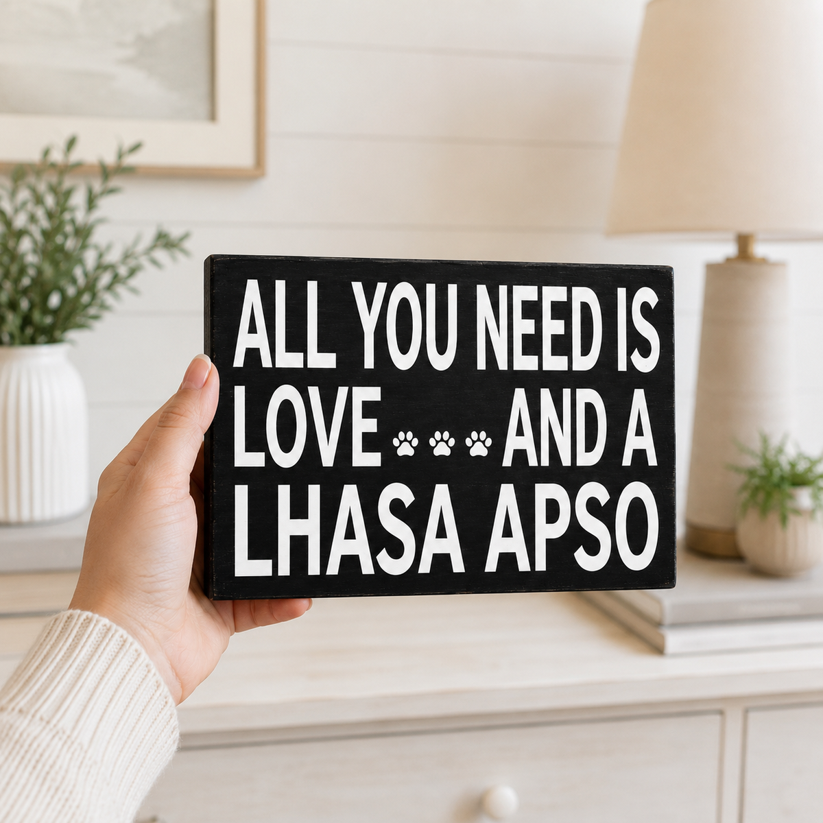 A hand holds a black rectangular wooden sign with bold white text that reads 'ALL YOU NEED IS LOVE AND A LHASA APSO' with three paw prints. The background shows a softly blurred home interior with a lamp, plants, and light-colored furniture.