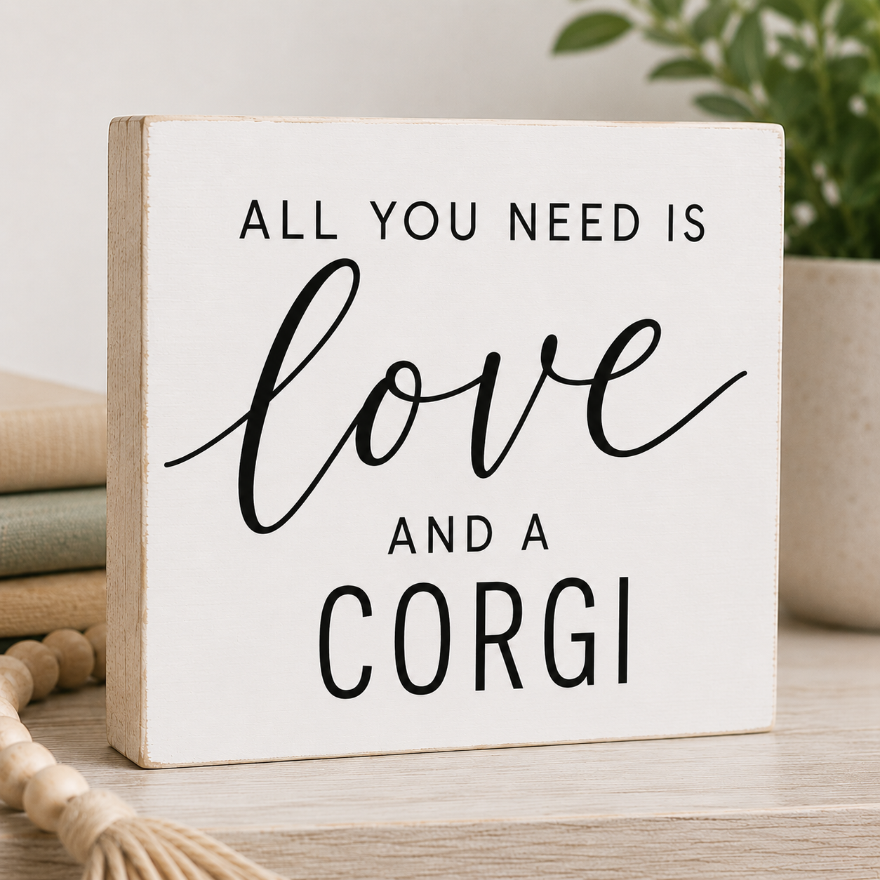 A square decorative sign with a white face and natural wood sides, printed with the phrase 'ALL YOU NEED IS love AND A CORGI' in black lettering, displayed on a light wood surface with neutral decor in the background.