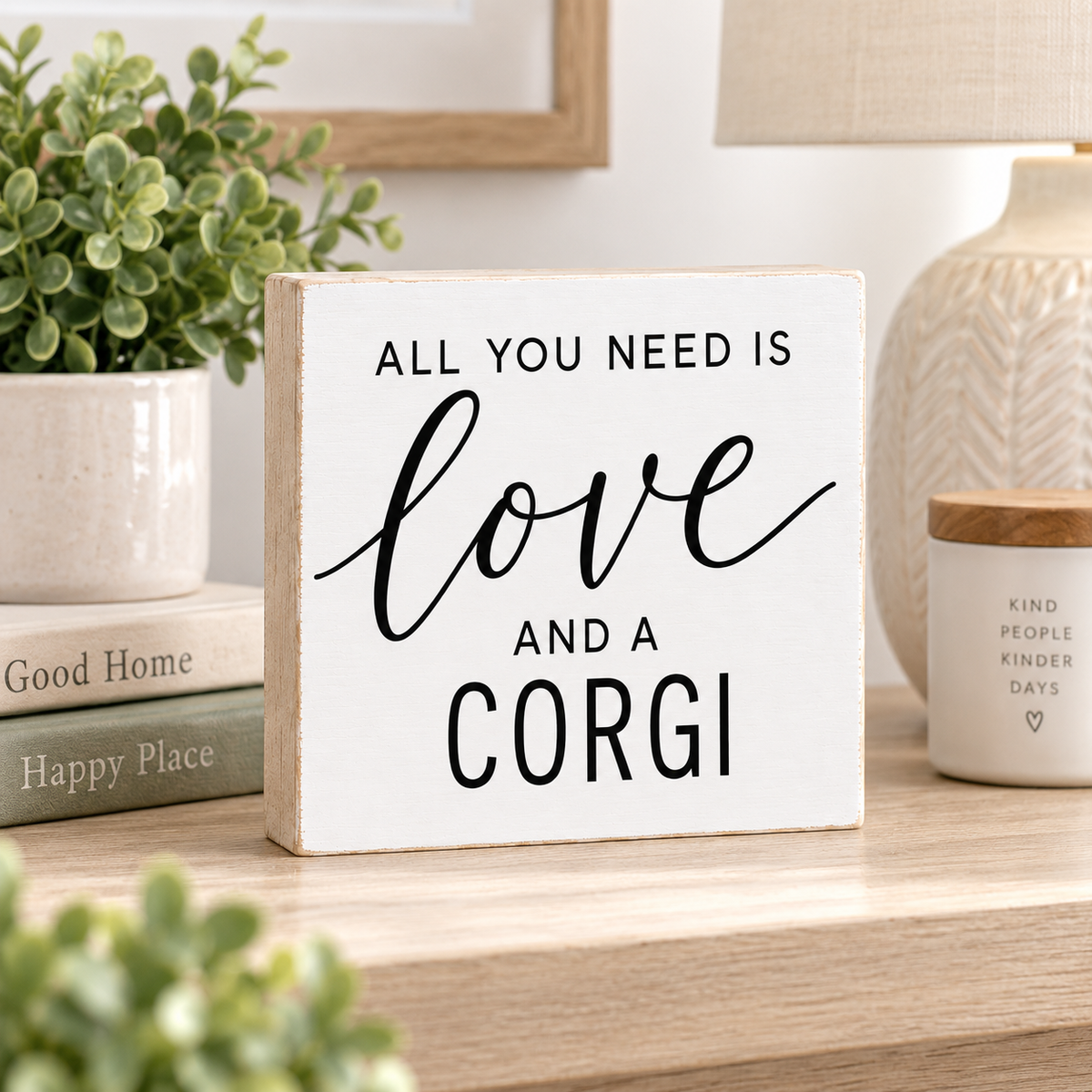 A square white decorative sign with black text reading 'ALL YOU NEED IS love AND A CORGI' sits on a wooden shelf in a styled home setting with a potted plant, books, and a candle.
