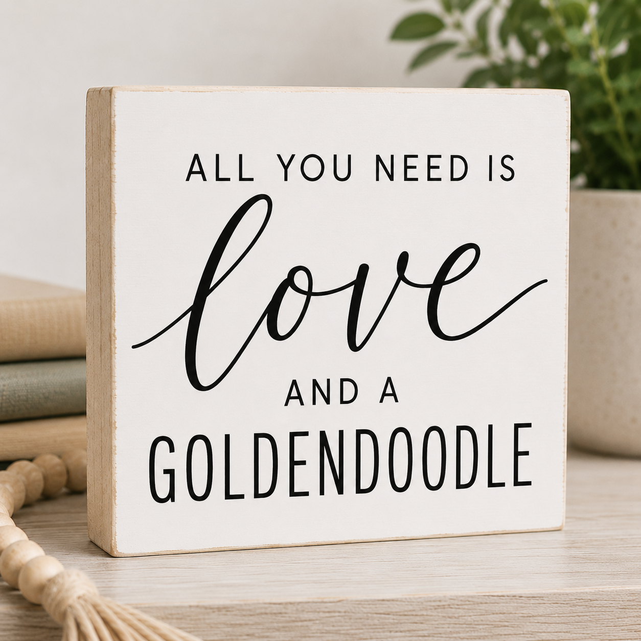A square white decorative wooden sign with black text reading 'ALL YOU NEED IS love AND A GOLDENDOODLE,' styled on a tabletop with books, a plant, and a tassel in the background.