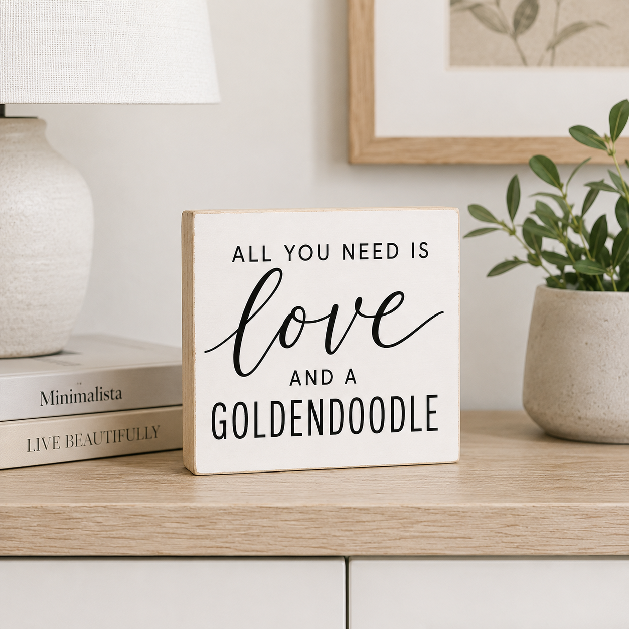 A square white decorative sign with a light wood border and black text reading 'ALL YOU NEED IS love AND A GOLDENDOODLE' sits on a light wood table in a styled room setting with a lamp, books, framed art, and a potted plant.