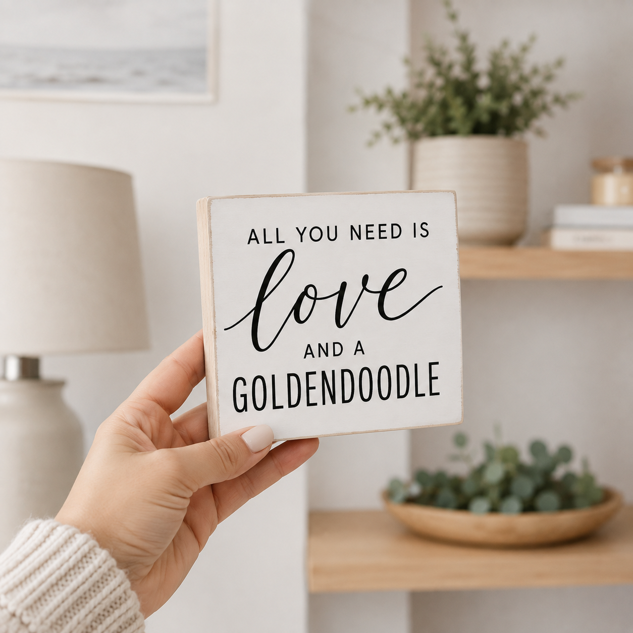A hand holds a square white wooden sign with black text that reads 'ALL YOU NEED IS love AND A GOLDENDOODE'. The background shows a softly blurred home interior with a lamp, shelves, and potted plants.