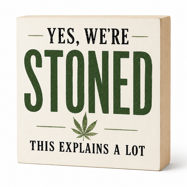 A square decorative wooden sign with a cream front and natural wood sides, printed with the phrase “YES, WE’RE STONED THIS EXPLAINS A LOT” in black and dark green lettering, plus a green leaf graphic, on a white background.
