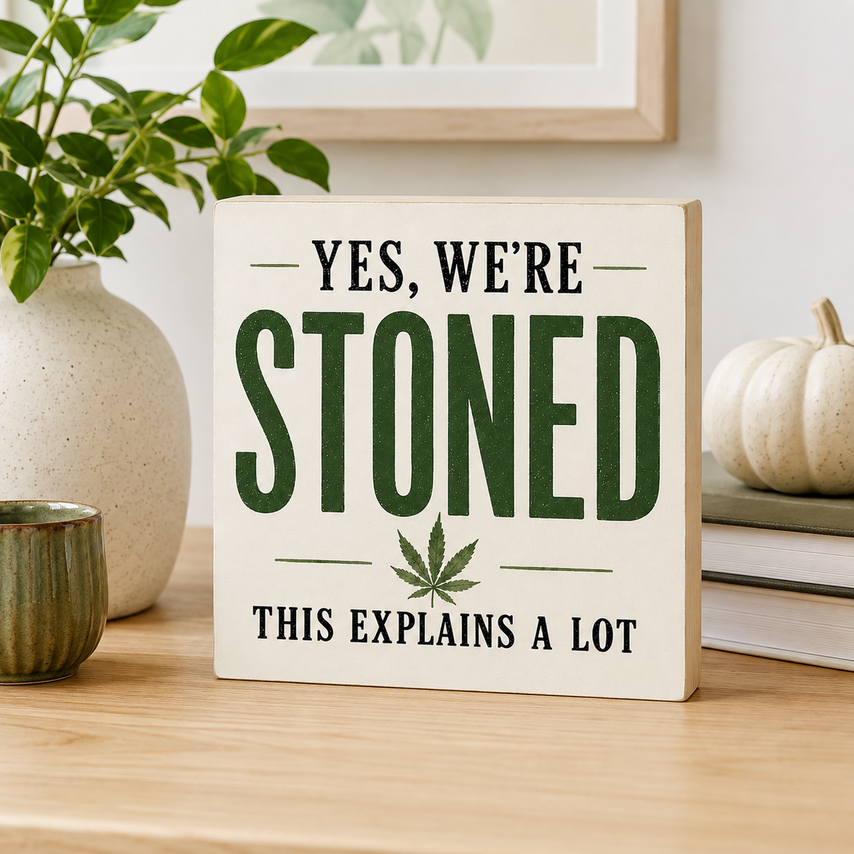 A square decorative sign on a wooden table reading “YES, WE’RE STONED THIS EXPLAINS A LOT” with large dark green text, black secondary text, and a cannabis leaf graphic. The sign is styled in a home setting with a plant in a vase, a small cup, books, and a small pumpkin in the background.