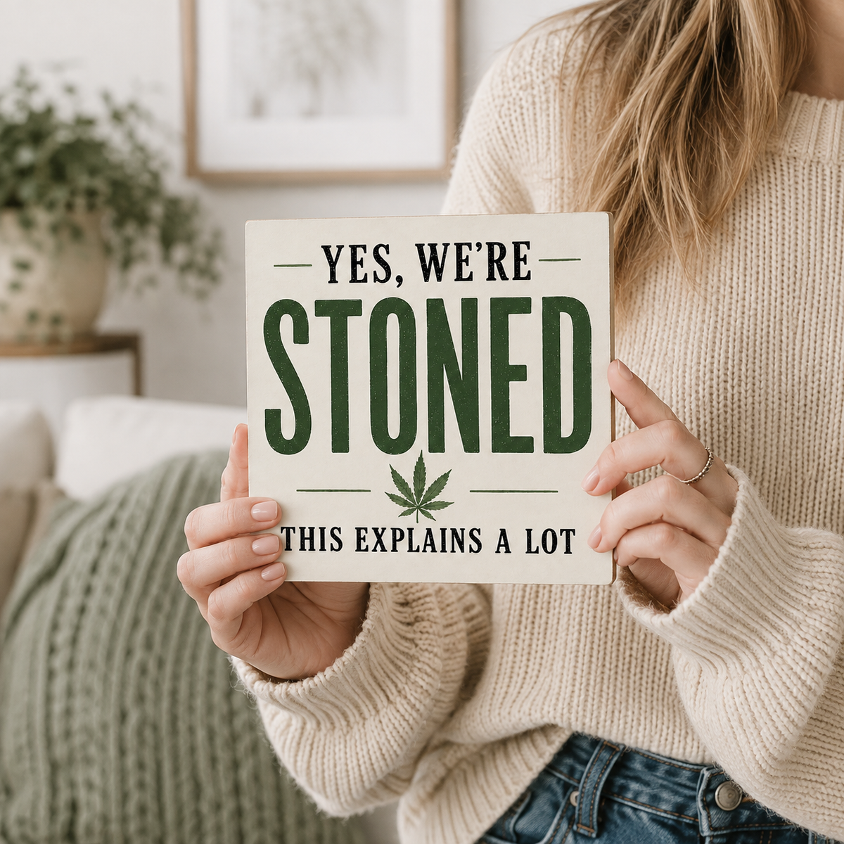 A person in a cream knit sweater holds a square sign that reads “YES, WE’RE STONED THIS EXPLAINS A LOT” with a green leaf graphic, photographed in a cozy indoor setting.