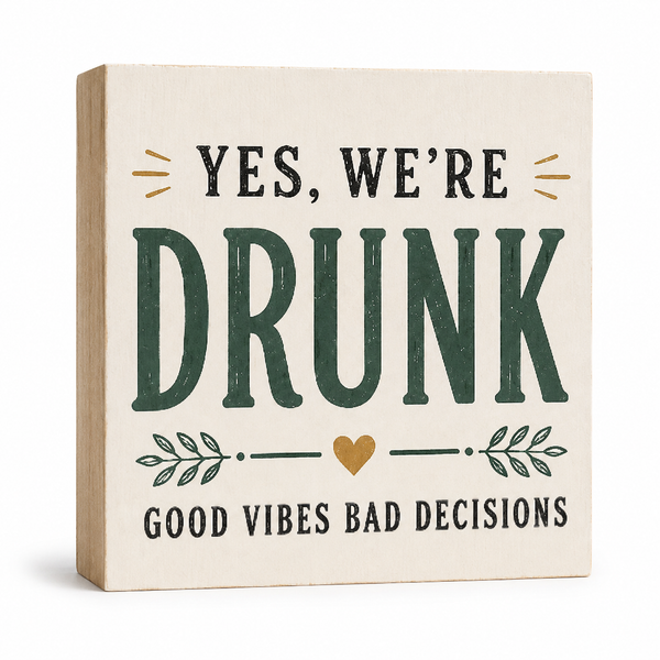 A square decorative wooden sign with a light beige wood-grain face and side, printed with the phrase 'YES, WE'RE DRUNK GOOD VIBES BAD DECISIONS' in dark green and black lettering, with a small gold heart and leaf motifs, shown on a white background.