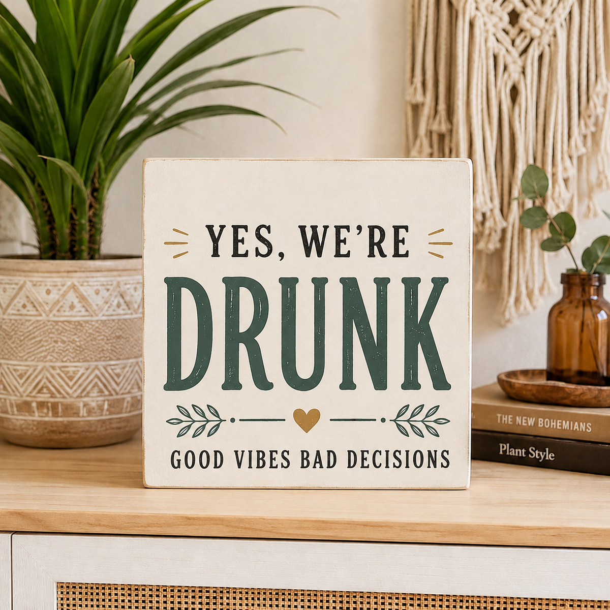A square wooden sign with a distressed cream background and dark green text reading 'YES, WE'RE DRUNK' and 'GOOD VIBES BAD DECISIONS,' styled on a tabletop with a potted plant, books, and a macrame wall hanging in the background.