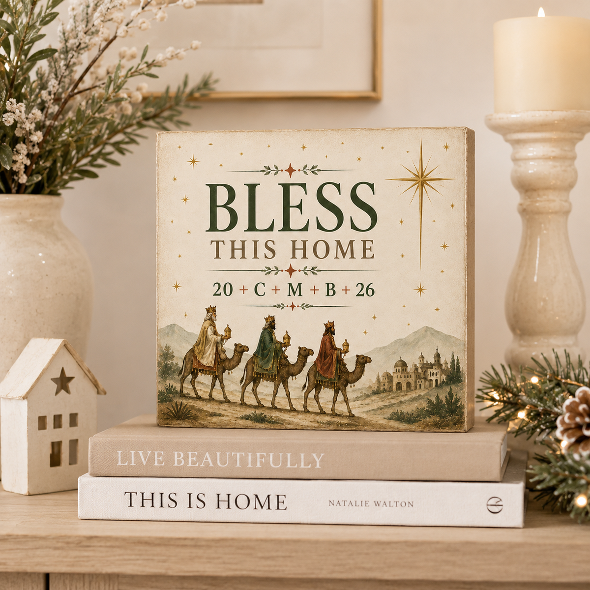 A decorative book titled 'Bless This Home' with a vintage-style illustration of three camels and stars, styled on top of two other books on a wooden shelf with holiday decor, a candle, and a small house ornament.