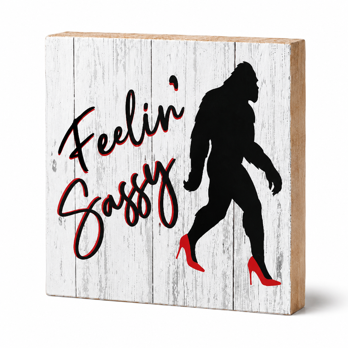 A square decorative wooden block sign with a whitewashed wood-look face, natural wood edges, the phrase 'Feelin' Sassy' in black script with red shadowing, and a black silhouette of Bigfoot wearing red high heels.