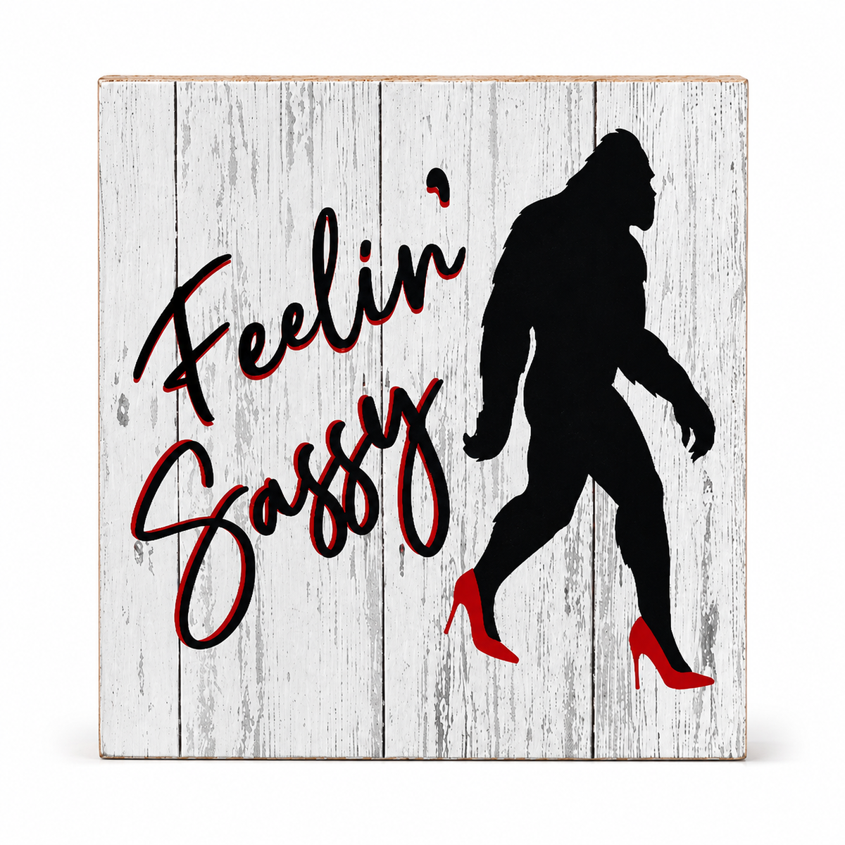 A square decorative wall sign with a distressed white wood background, featuring the phrase 'Feelin' Sassy' in black script with red outline and a black silhouette of a Bigfoot figure wearing red high heels.