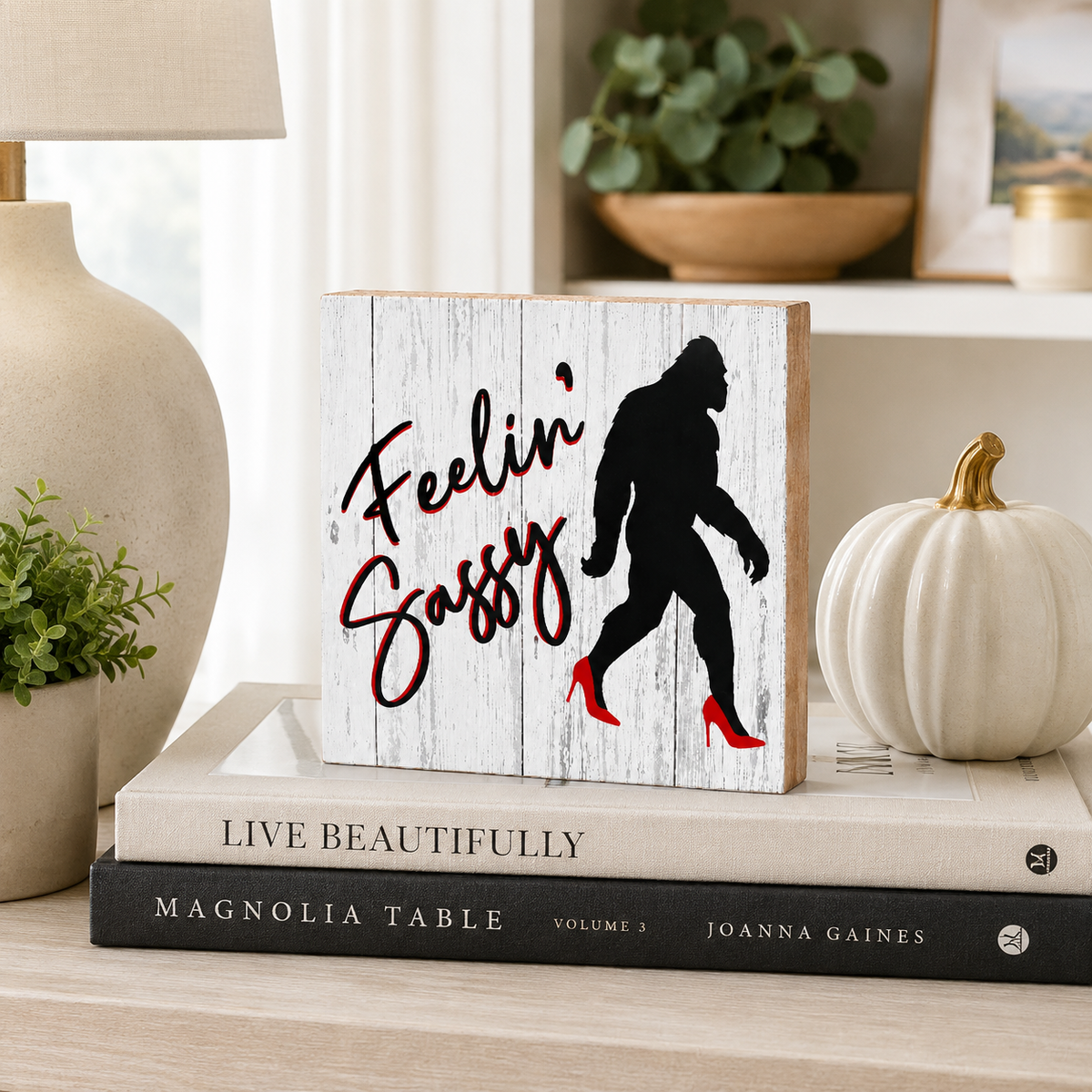 A decorative wooden sign with a distressed white wood background, featuring the phrase 'Feelin' Sassy' in red script and a black silhouette of a Bigfoot-like figure walking in red high heels. The sign is styled on top of two books on a light wood table, surrounded by a beige ceramic lamp, a small potted plant, and a white decorative pumpkin.