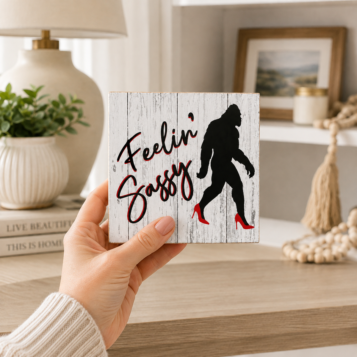 A hand holds a square white wood-look sign with the phrase “Feelin’ Sassy” in black script and a black Bigfoot silhouette wearing red high heels, shown in a softly lit home interior.