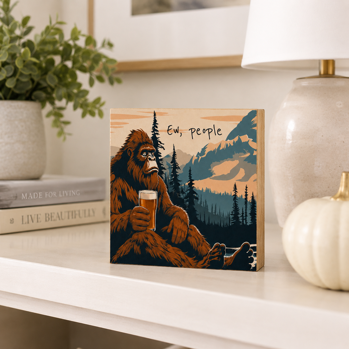 A square wooden art print or block featuring a retro-style illustration of a big cat-like figure holding a drink in front of mountains and trees, with the text 'Ev, people.' It is displayed on a white shelf in a styled home setting with a plant, books, lamp, and pumpkin.