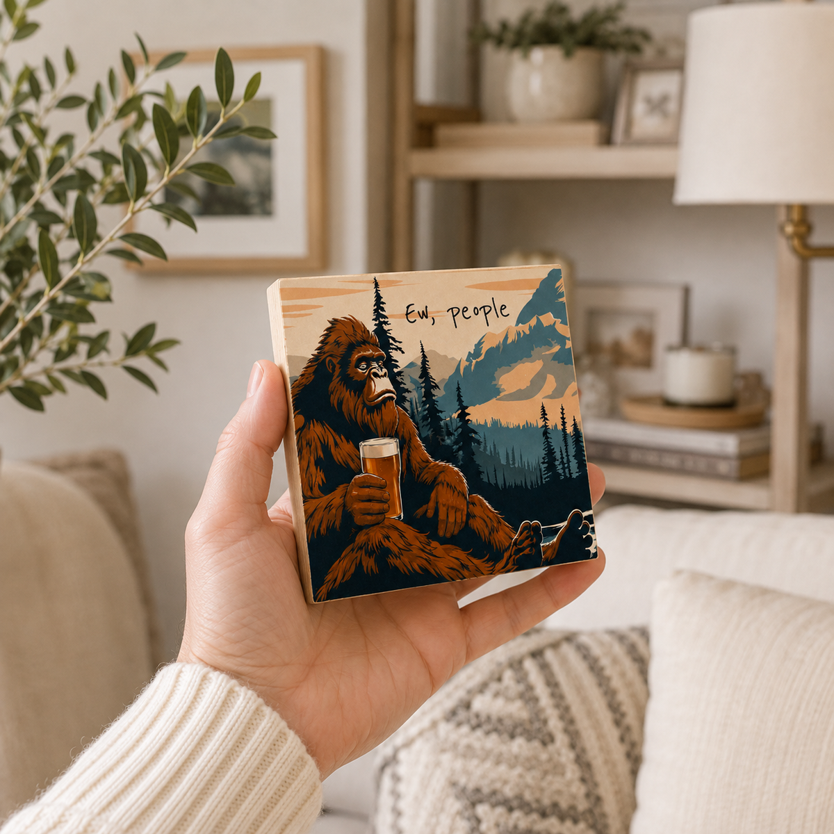A hand holds a square wooden coaster featuring a retro illustration of a Bigfoot sitting in a forest with mountains and the text 'Ev, people'. The background shows a cozy, softly lit living room with a lamp, shelves, and cushions.