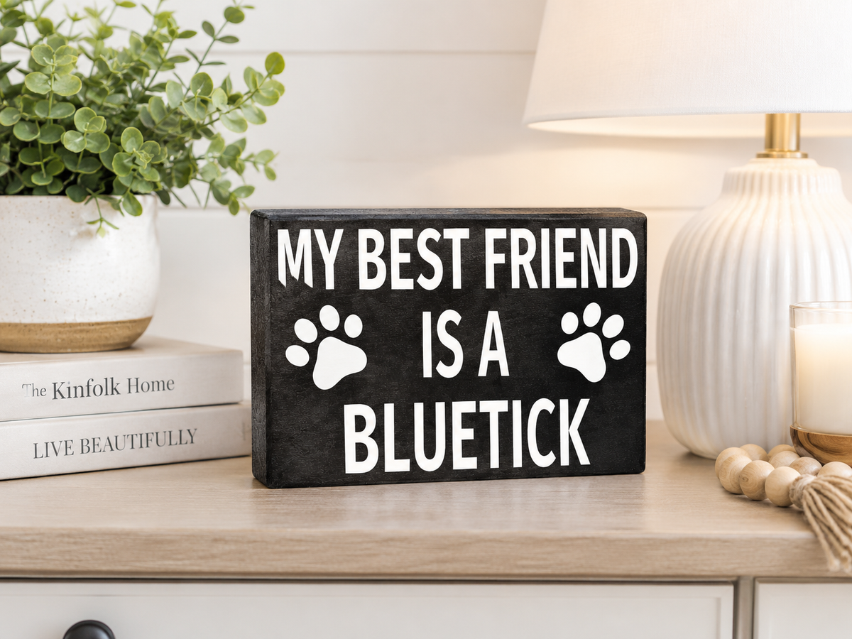 A rectangular black wooden sign with white text reading 'MY BEST FRIEND IS A BLUETICK' and two white paw prints, displayed on a light wood table in a styled home setting with a lamp, candle, books, and a plant.
