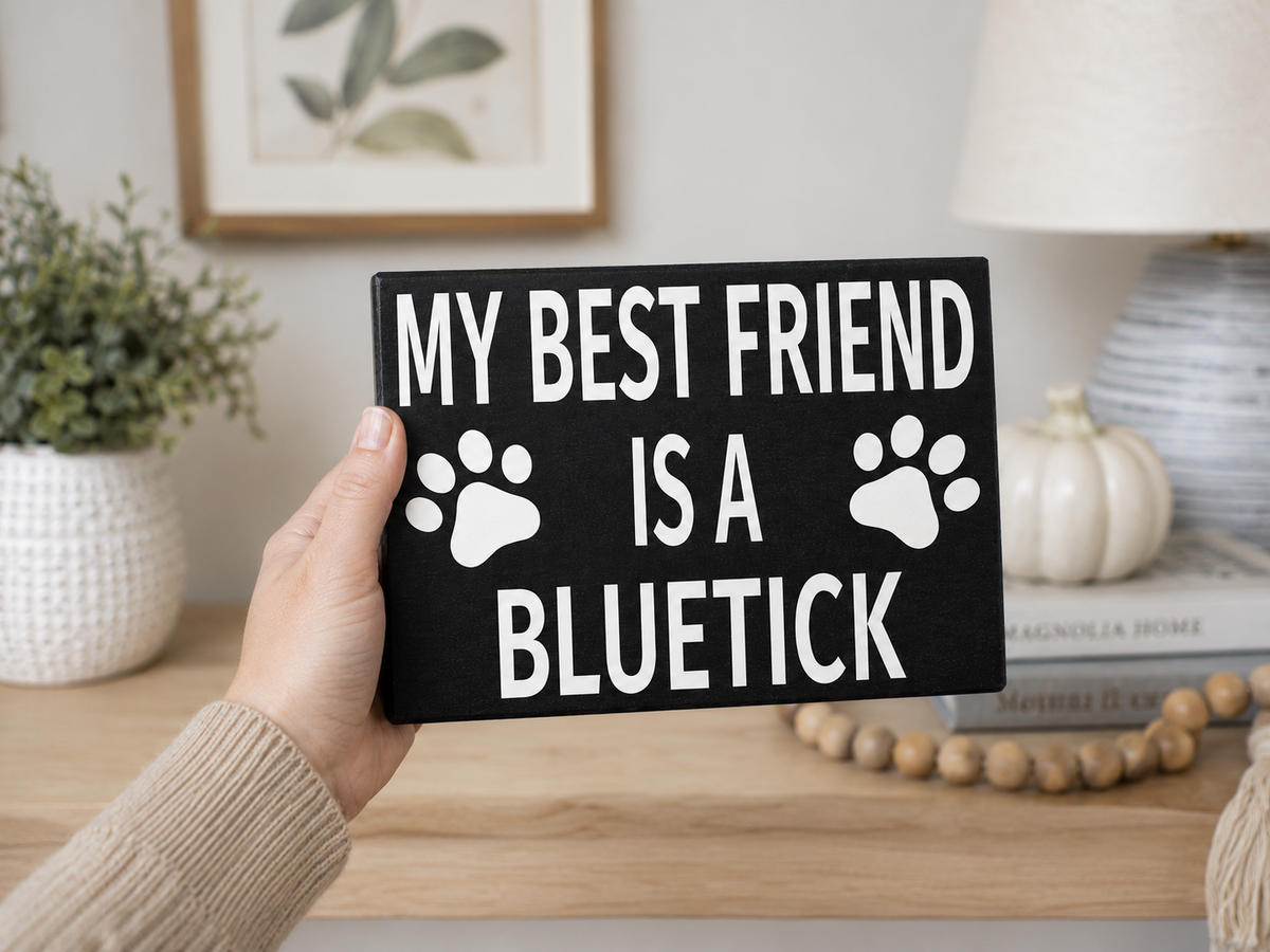 A hand holds a black rectangular sign with white text reading 'MY BEST FRIEND IS A BLUETICK' and two white paw prints. The background shows a cozy indoor setting with a wooden shelf, books, a lamp, and a small pumpkin.