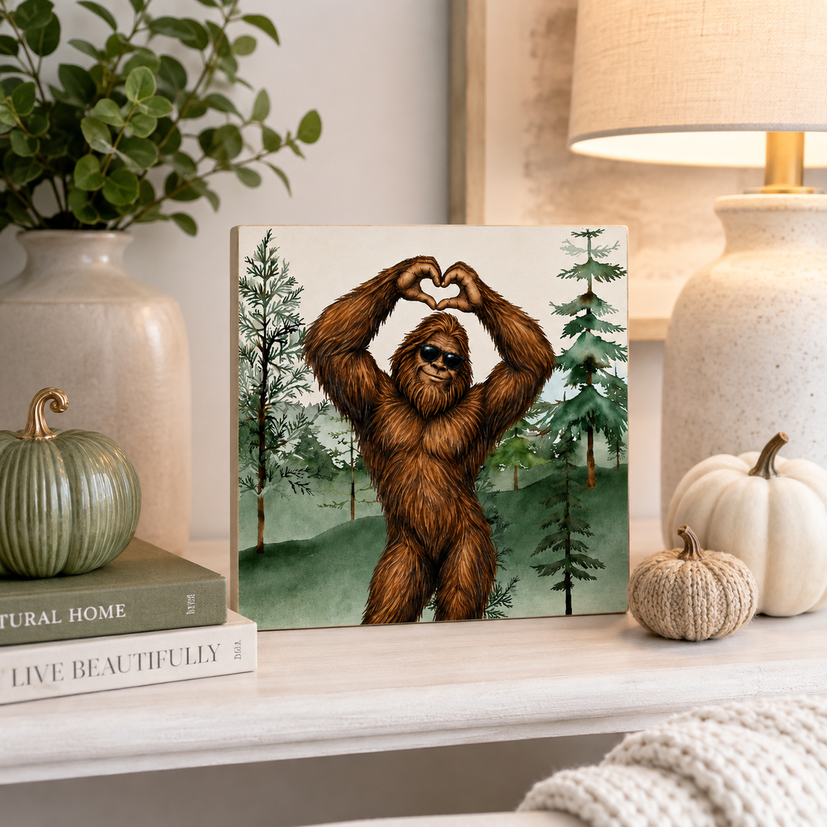 A square art print featuring a brown furry creature forming a heart with its hands, set against a forest background, displayed on a shelf in a styled home decor setting.