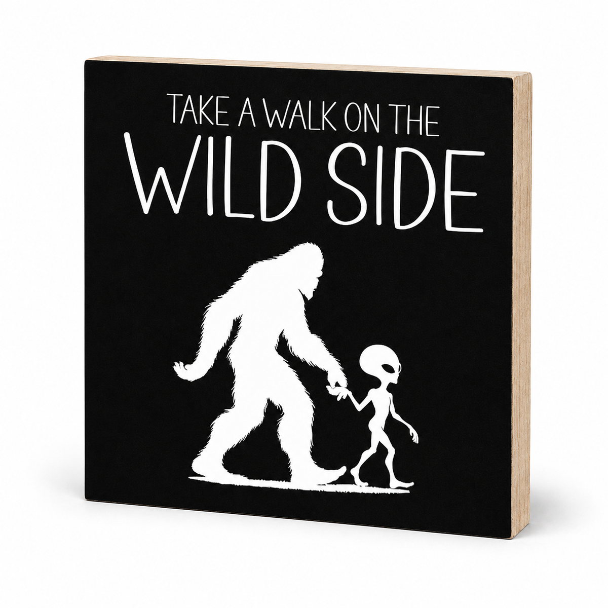 A square black decorative sign with a natural wood side edge, featuring the white text 'TAKE A WALK ON THE WILD SIDE' above a white silhouette of a Bigfoot figure holding hands with a small alien.