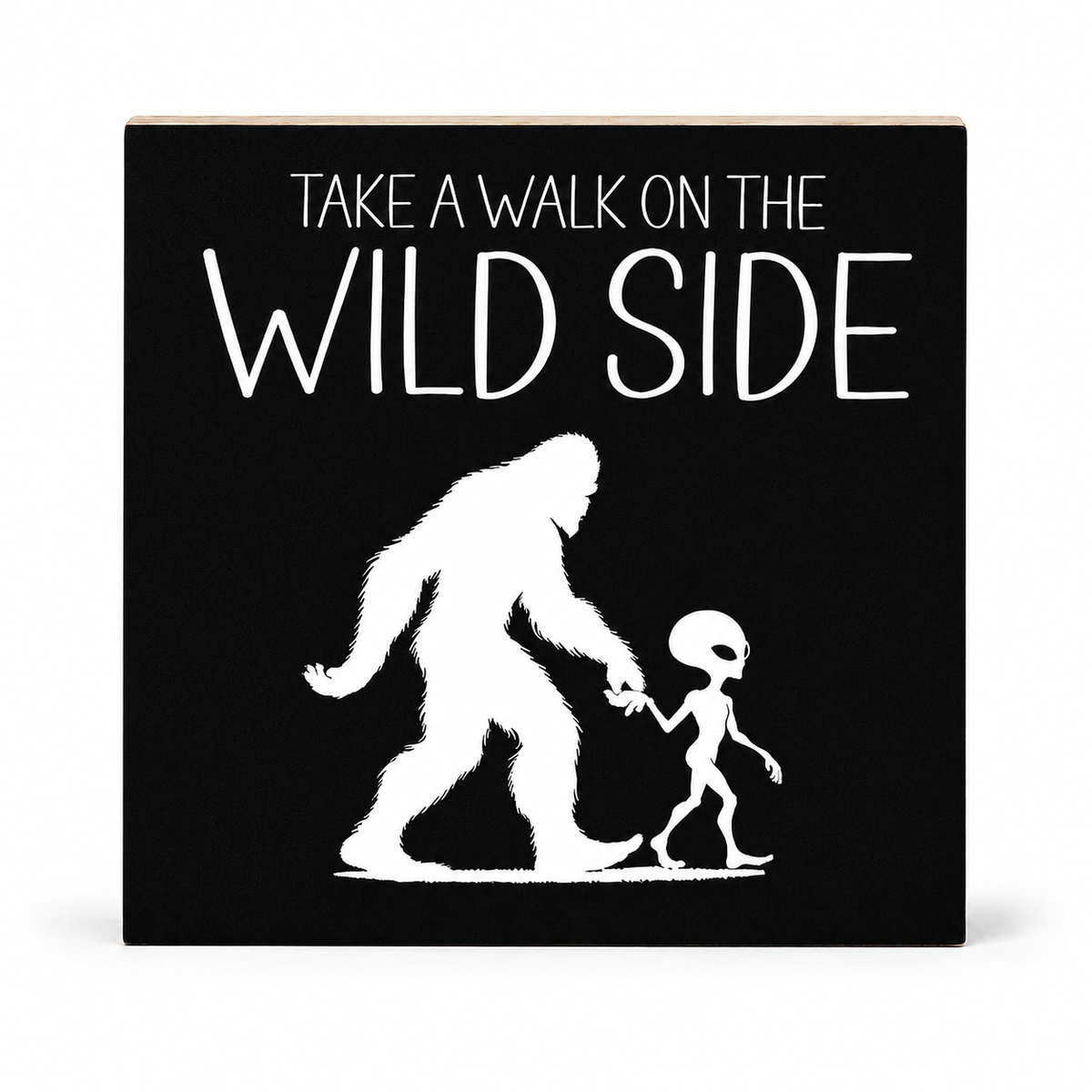A square black decorative sign with a light wood edge, featuring the white text 'TAKE A WALK ON THE WILD SIDE' above a white silhouette of a Bigfoot-like figure holding hands with an alien, shown on a white background.