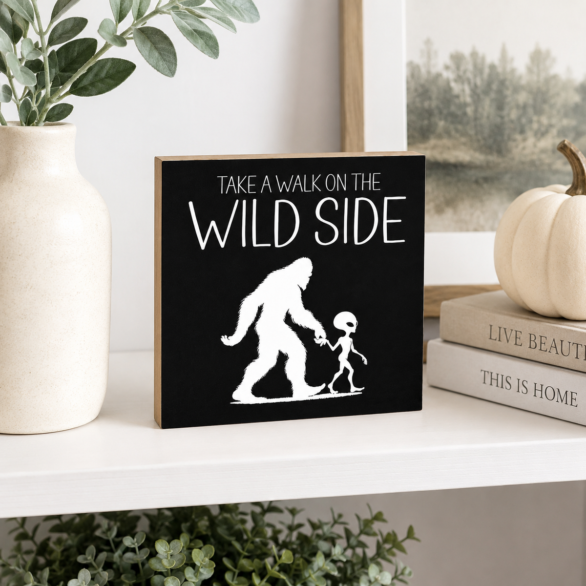 A square black decorative sign with white text reading 'TAKE A WALK ON THE WILD SIDE' and a white silhouette of a big-footed figure holding hands with a small figure, displayed on a white shelf with a vase, framed print, books, and a pumpkin around it.
