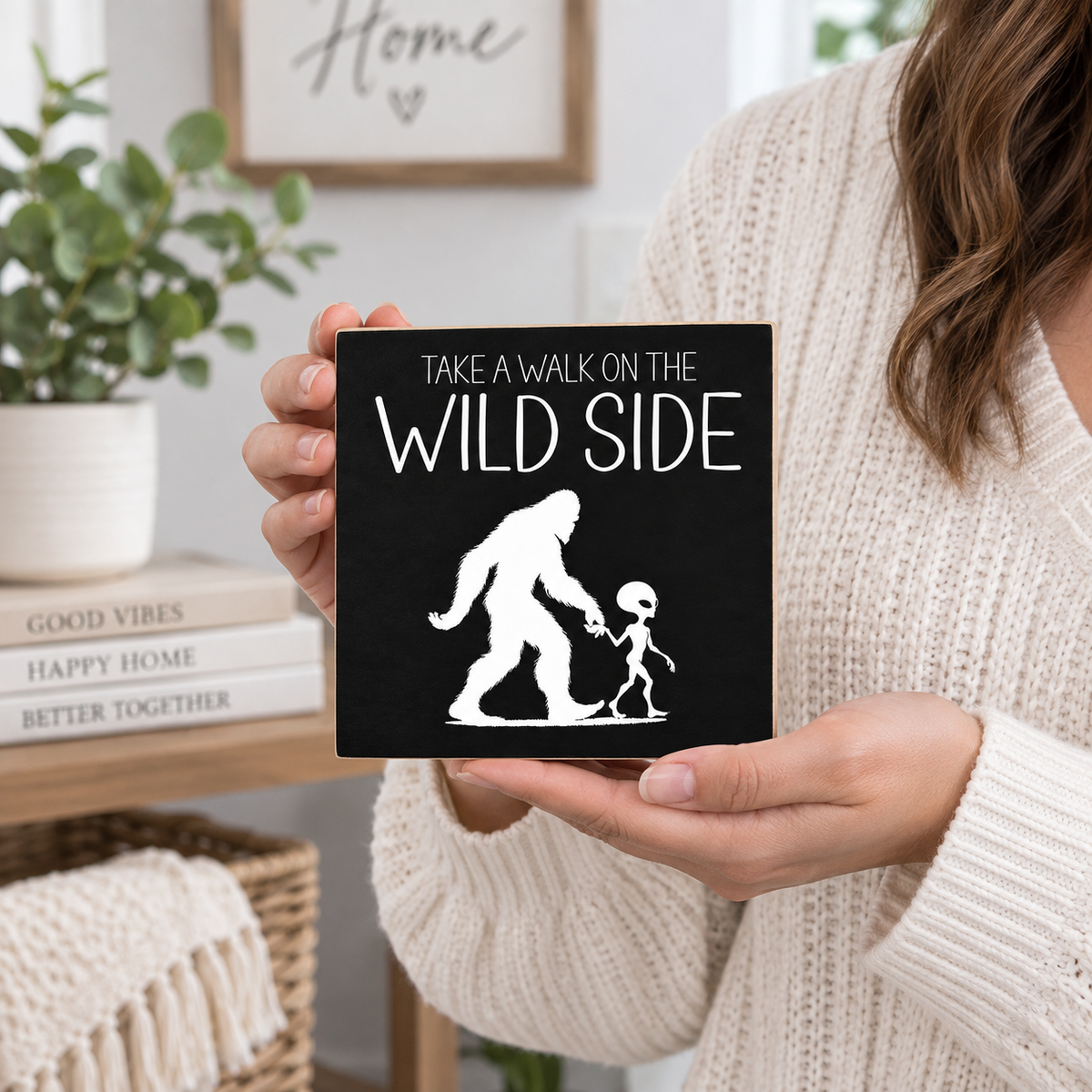 A person in a cream knit sweater holds a square black sign with white text reading 'TAKE A WALK ON THE WILD SIDE' and a white silhouette of a Bigfoot-like figure holding a child's hand. The background shows a softly blurred home interior with a plant, books, and decor.