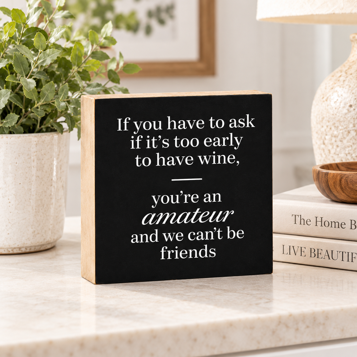 A square wooden sign with a black face and natural wood edges, featuring white text that reads, 'If you have to ask if it's too early to have wine, you're an amateur and we can't be friends.' It is styled on a marble countertop with a potted plant, books, and a lamp in the background.